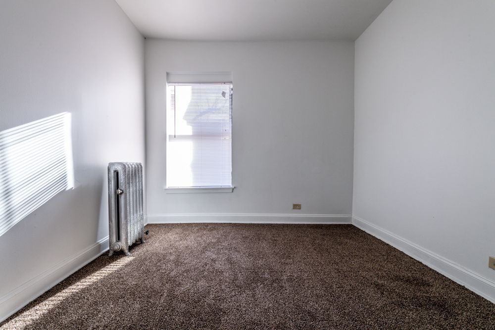 Empty room with white walls, brown carpet, window with blinds, and a radiator.