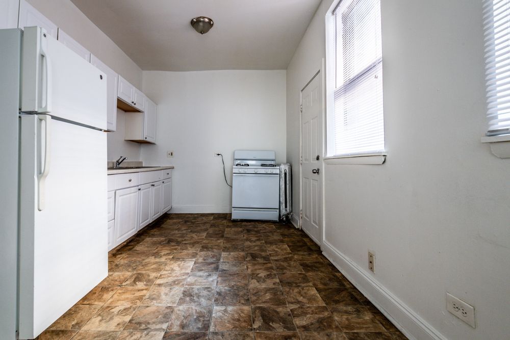 Small, white kitchen with refrigerator, cabinets, stove, door and window. Brown floor.