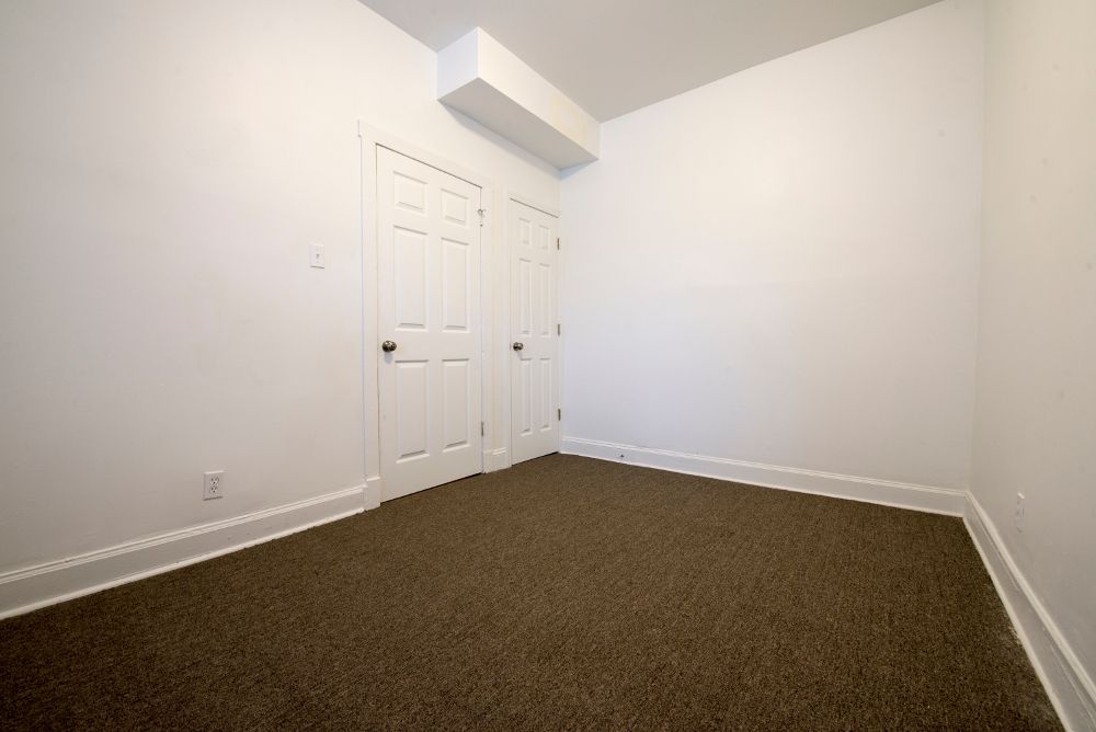 Empty room with brown carpet, white walls, two white doors, and a white ceiling beam.