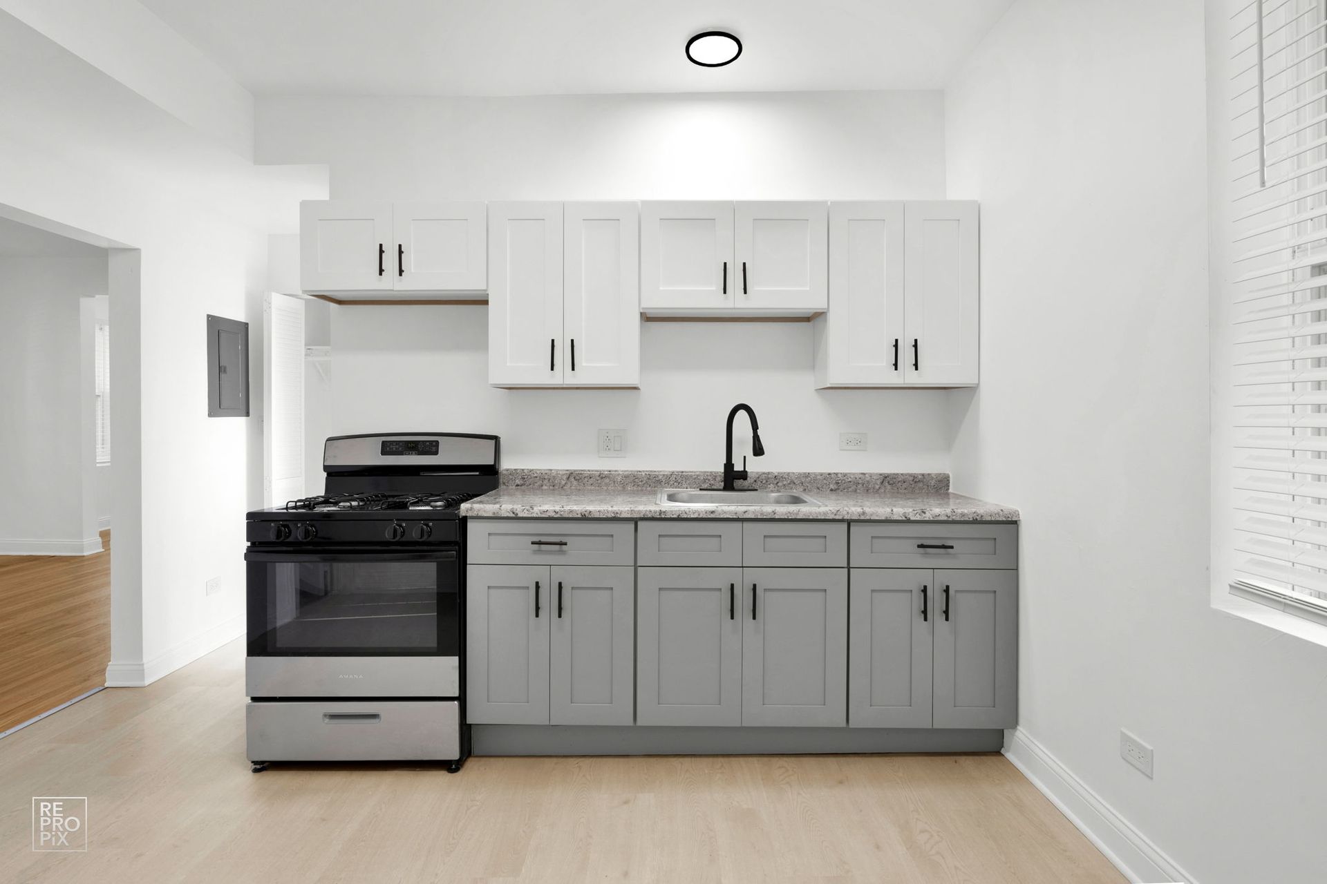 A kitchen with gray and white cabinets, black faucet, stove, and countertops. Room has wood floors and a window with blinds.