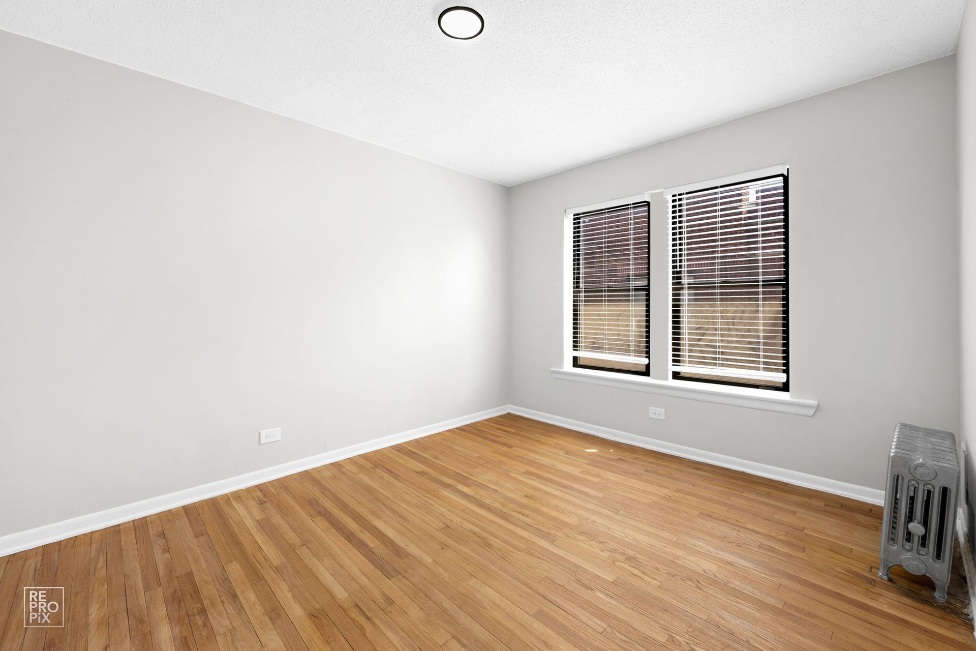 Empty room with hardwood floors, two windows with blinds, and a radiator.