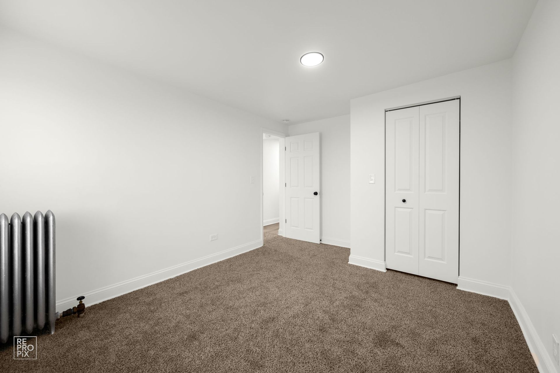 Empty room with white walls, brown carpet, a closet, and a radiator. A doorway leads to another room.