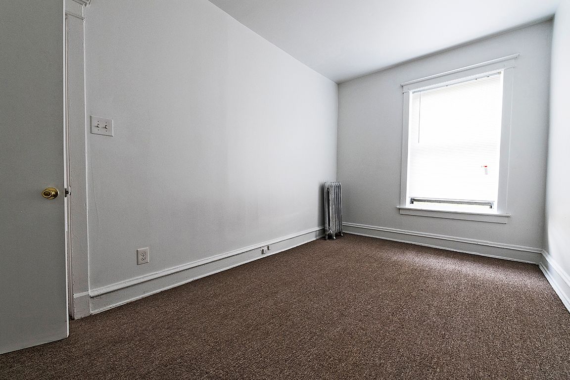 Empty room with neutral-colored walls and carpet. Window provides natural light. Door on left.