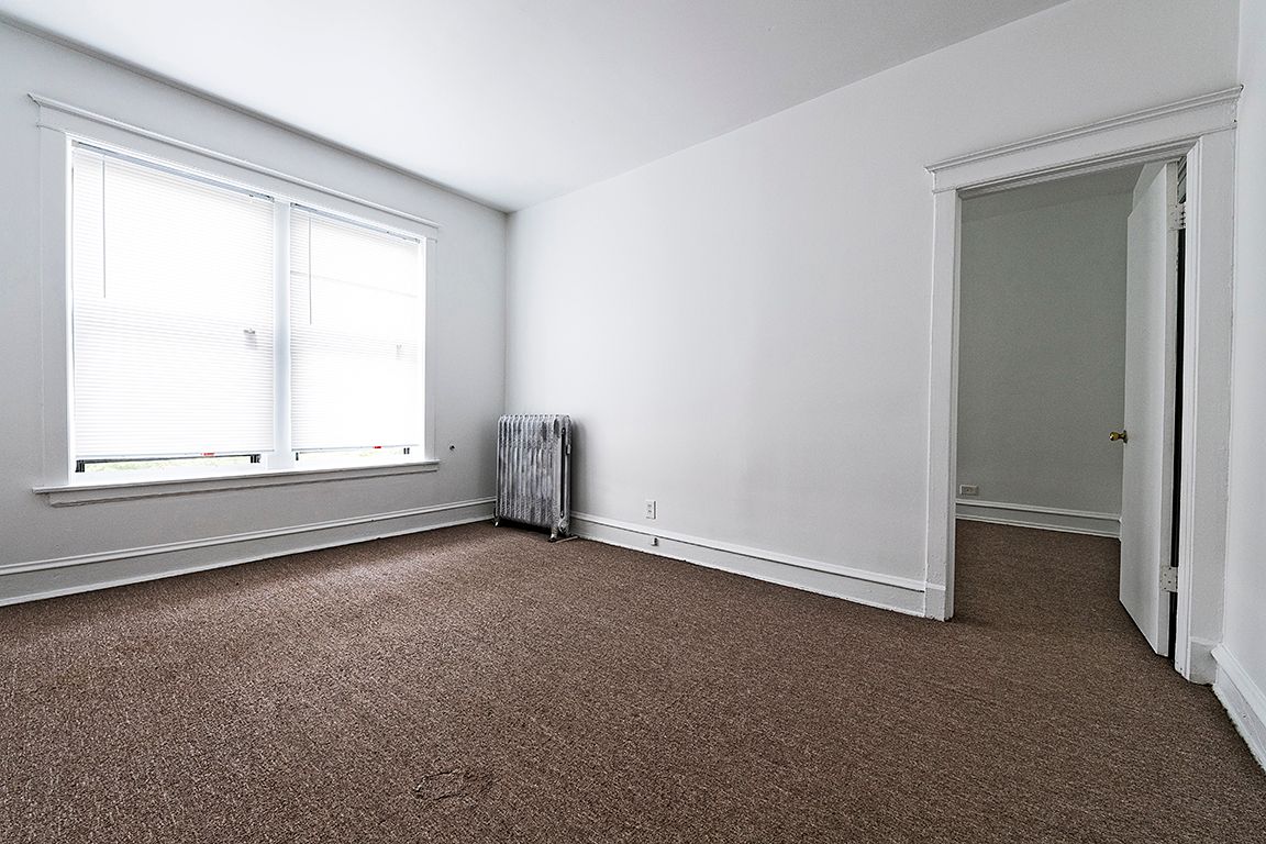 Empty room with brown carpet, white walls, window, radiator, and doorway.