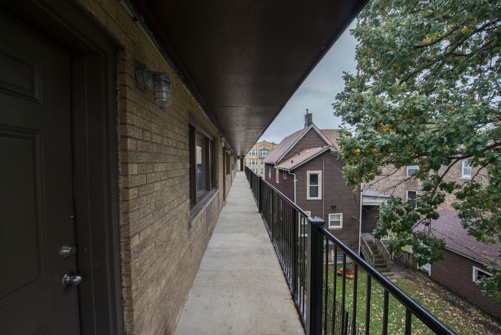 Exterior apartment hallway with brick wall and black railing overlooking backyard.