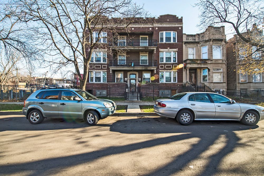 Cars parked on a street in front of a three-story brick apartment building. Bare trees and sunny day.