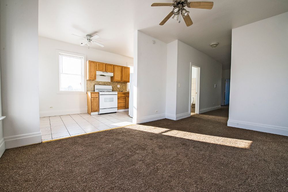 Bright, empty apartment interior with brown carpet, white walls, and a view into the kitchen.