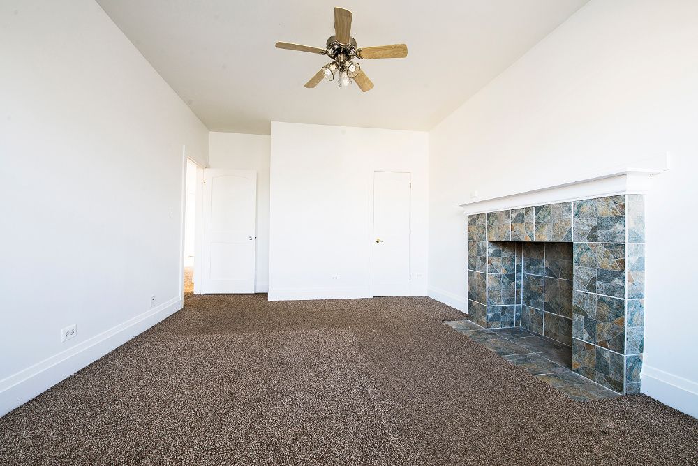 Empty room with brown carpet, white walls, and a tiled fireplace. Ceiling fan is visible.