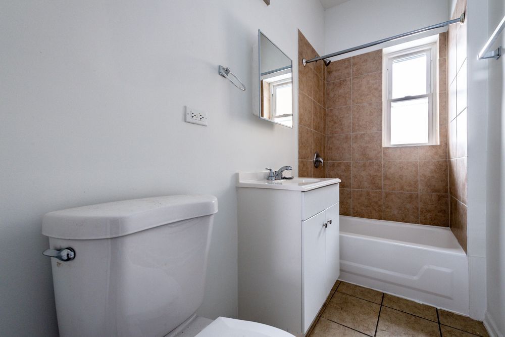 Bathroom with white toilet, vanity, and bathtub. Brown tile in shower area, window with natural light.