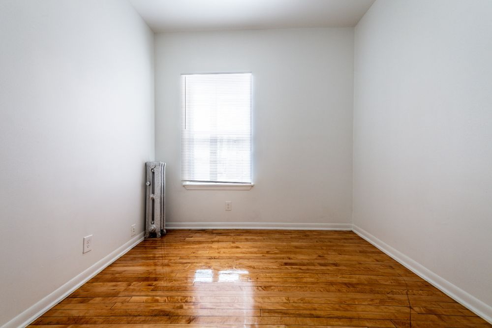 Empty room with hardwood floors, window with blinds, and a radiator against the wall. White walls.
