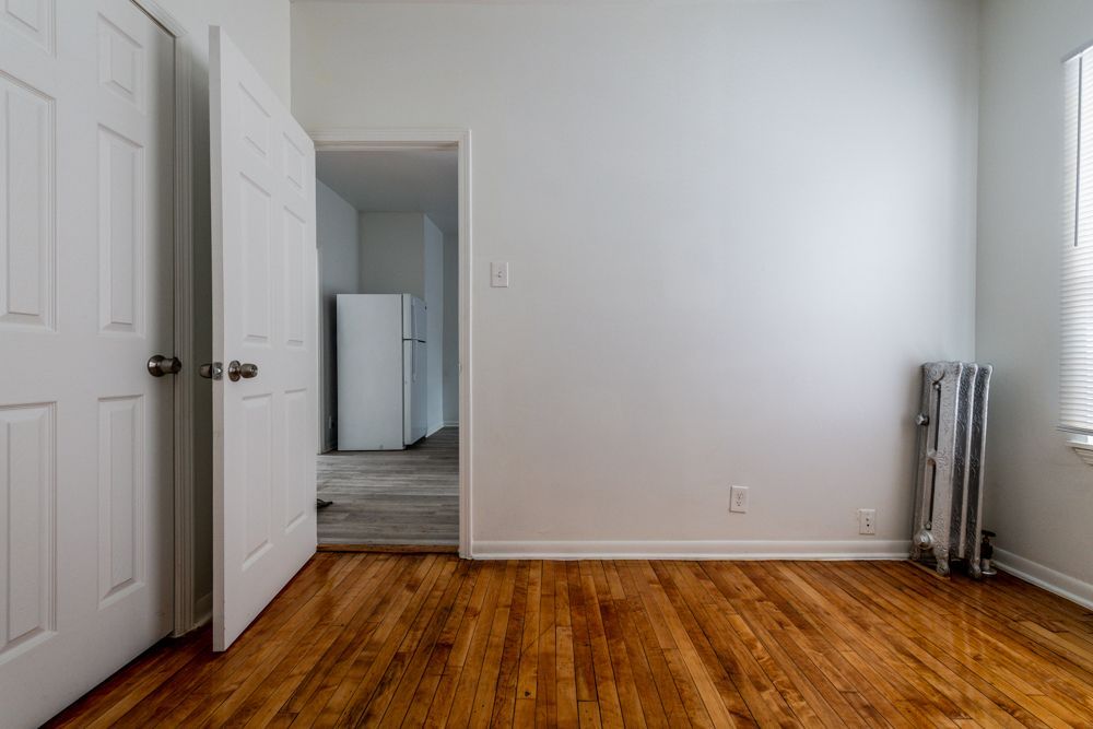 Empty room with hardwood floors, white walls, and a doorway leading to a room with a refrigerator.
