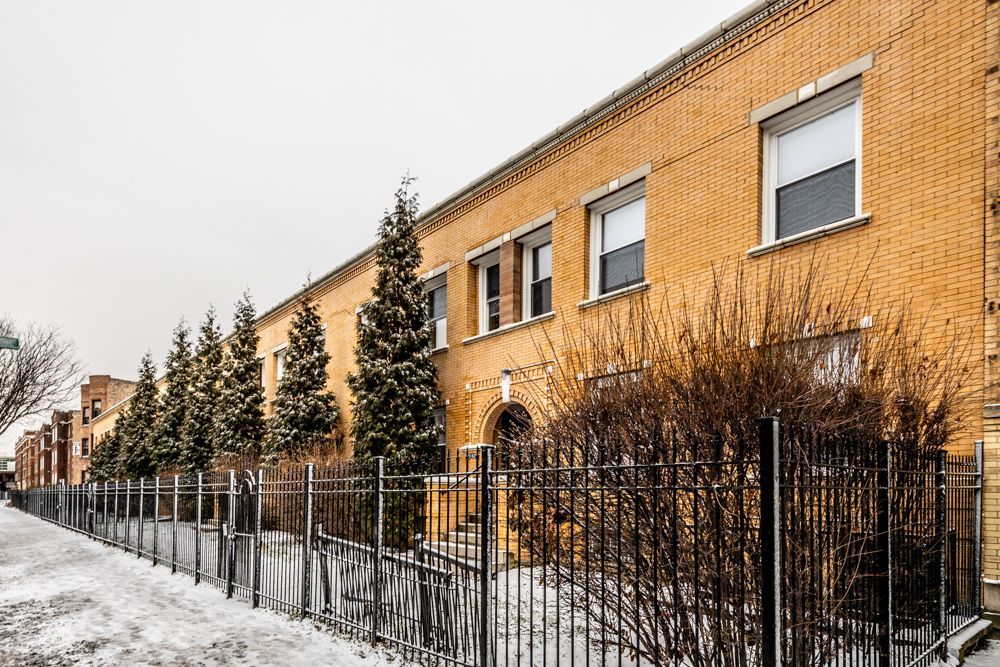 Brick building with snow-covered sidewalk and black metal fence. Evergreen trees line the path. Overcast day.