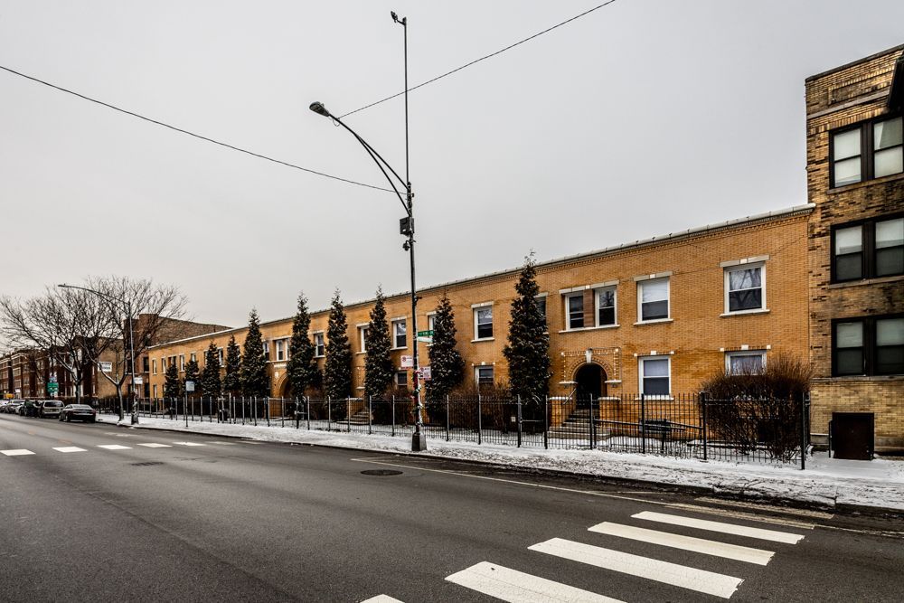 Apartment building on a street with snow. Brown brick, row of windows, trees. Cloudy sky.