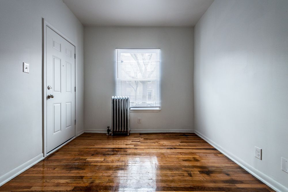 Empty room with a wooden floor, white walls, a window with blinds, and a closed door.
