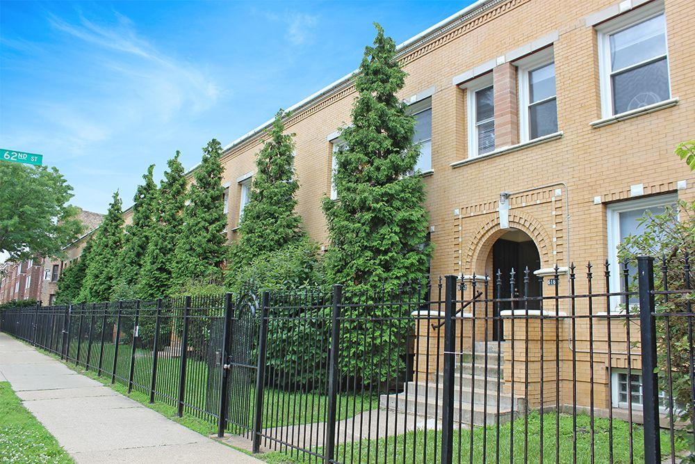 Row of brick townhomes behind a black metal fence, with green trees along the front and a sidewalk.