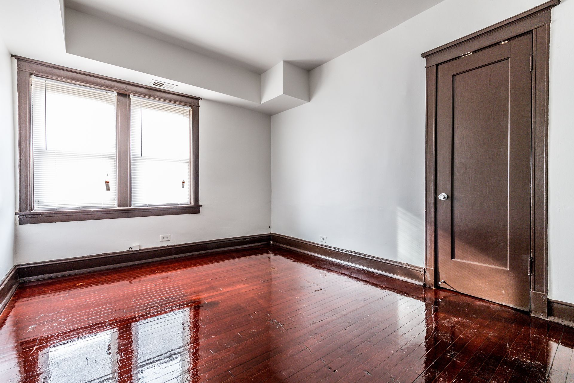 Empty room with hardwood floors, a window, and a closed brown door.