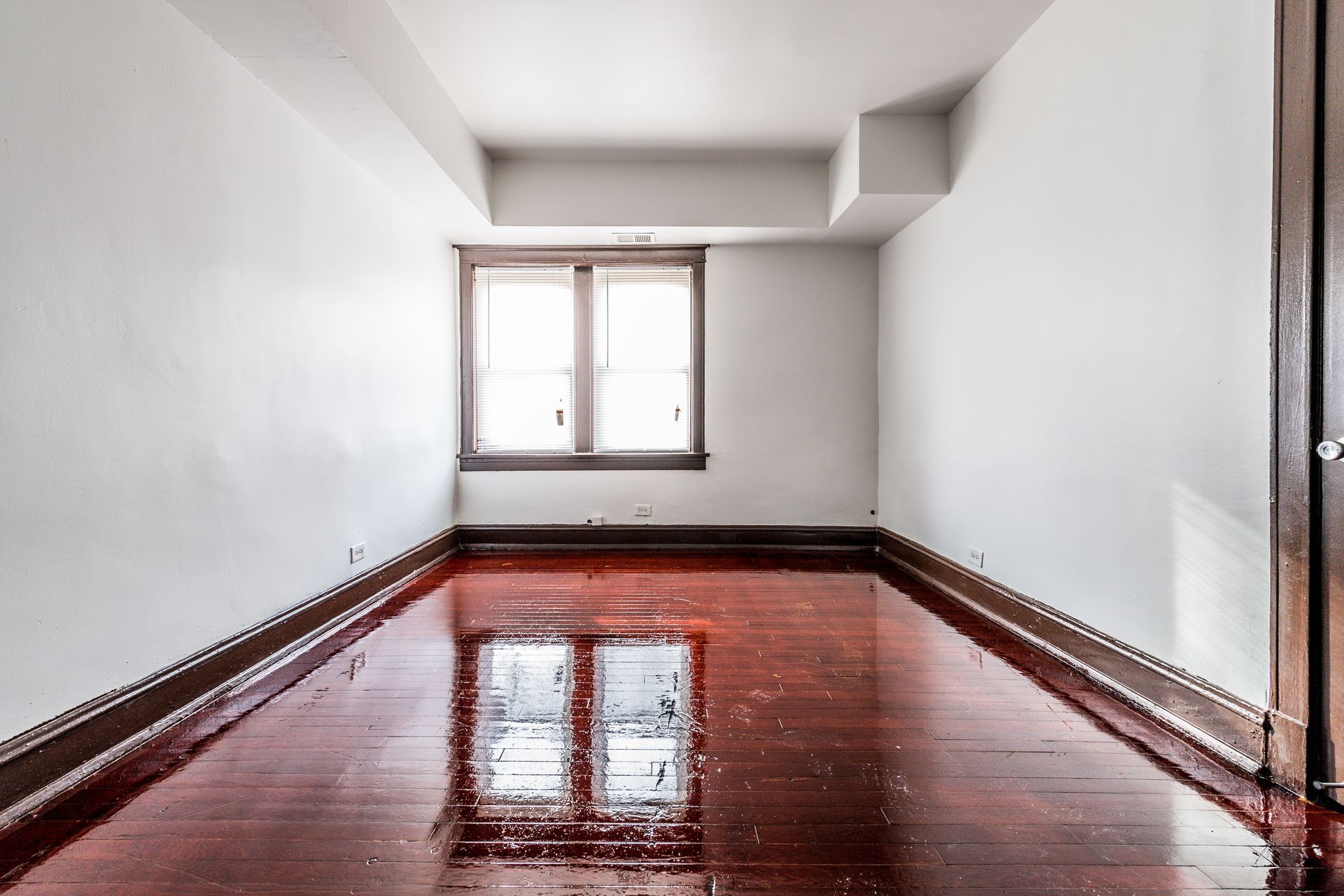 Empty room with white walls, window, and glossy red-brown wooden floor reflecting the window.