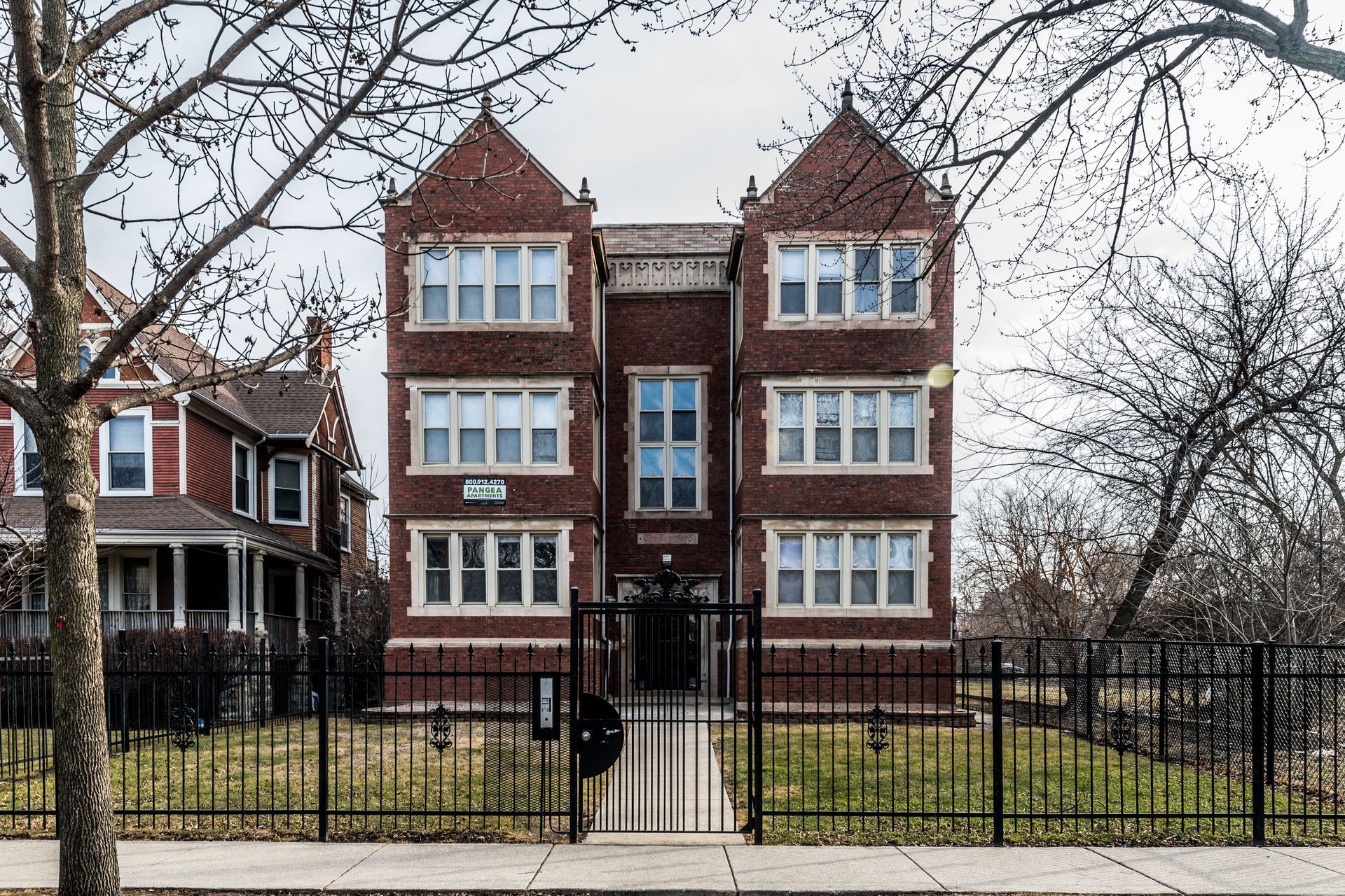 Three-story brick apartment building behind a black metal fence, flanked by bare trees and houses on a cloudy day.
