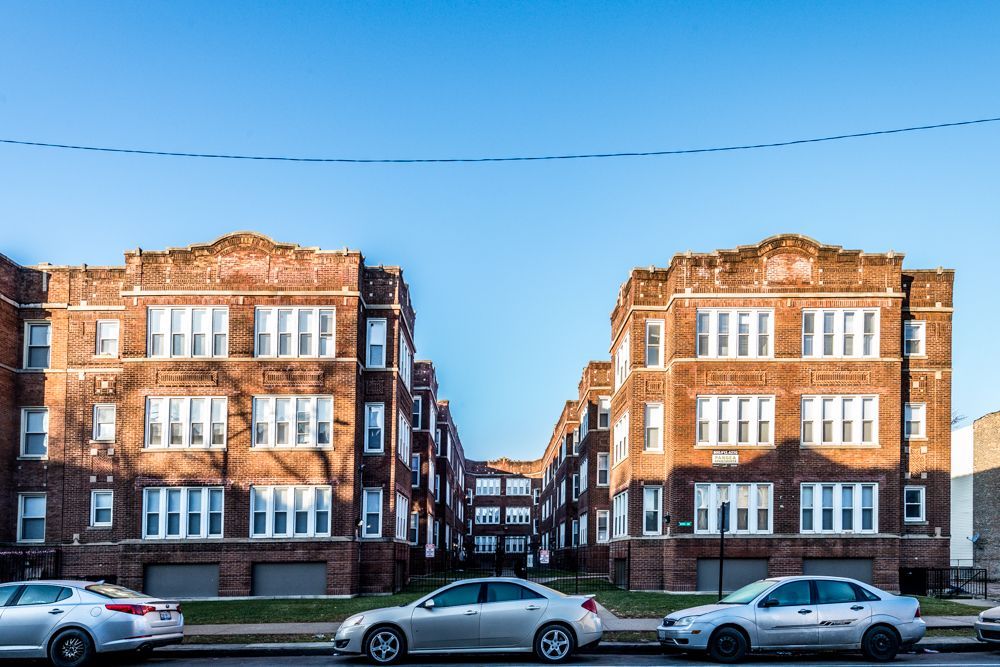 Red brick apartment buildings with cars parked in front under a clear blue sky.