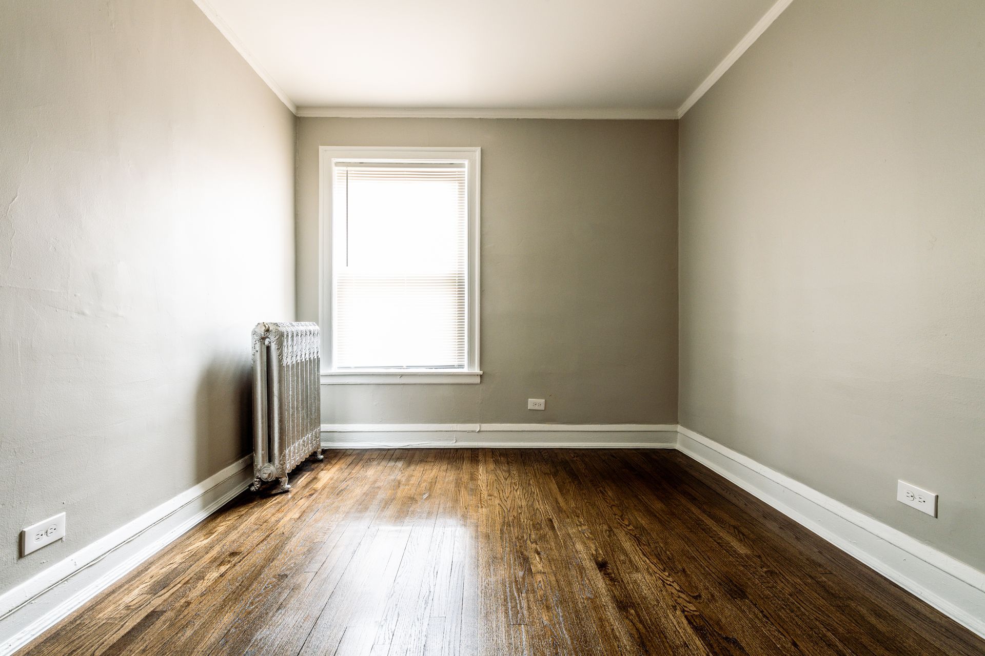 Empty room with wood floor, gray walls, radiator, and window with blinds.