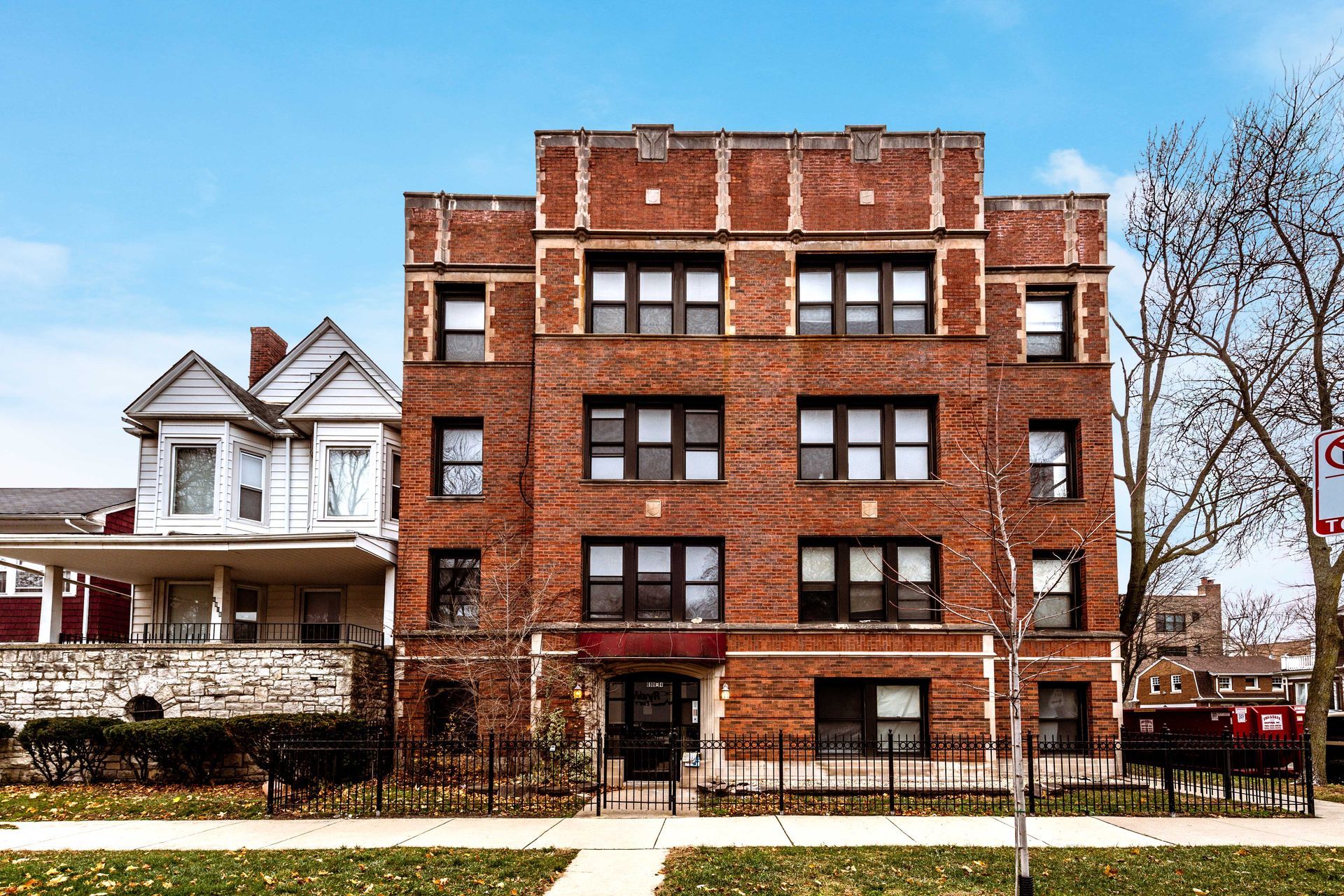 Red brick apartment building with black windows, fronted by a low iron fence, on a sunny day.