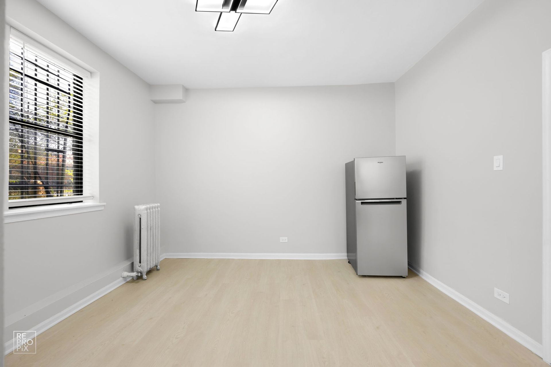 Empty, white-walled room with window, radiator, refrigerator, and light-colored flooring.