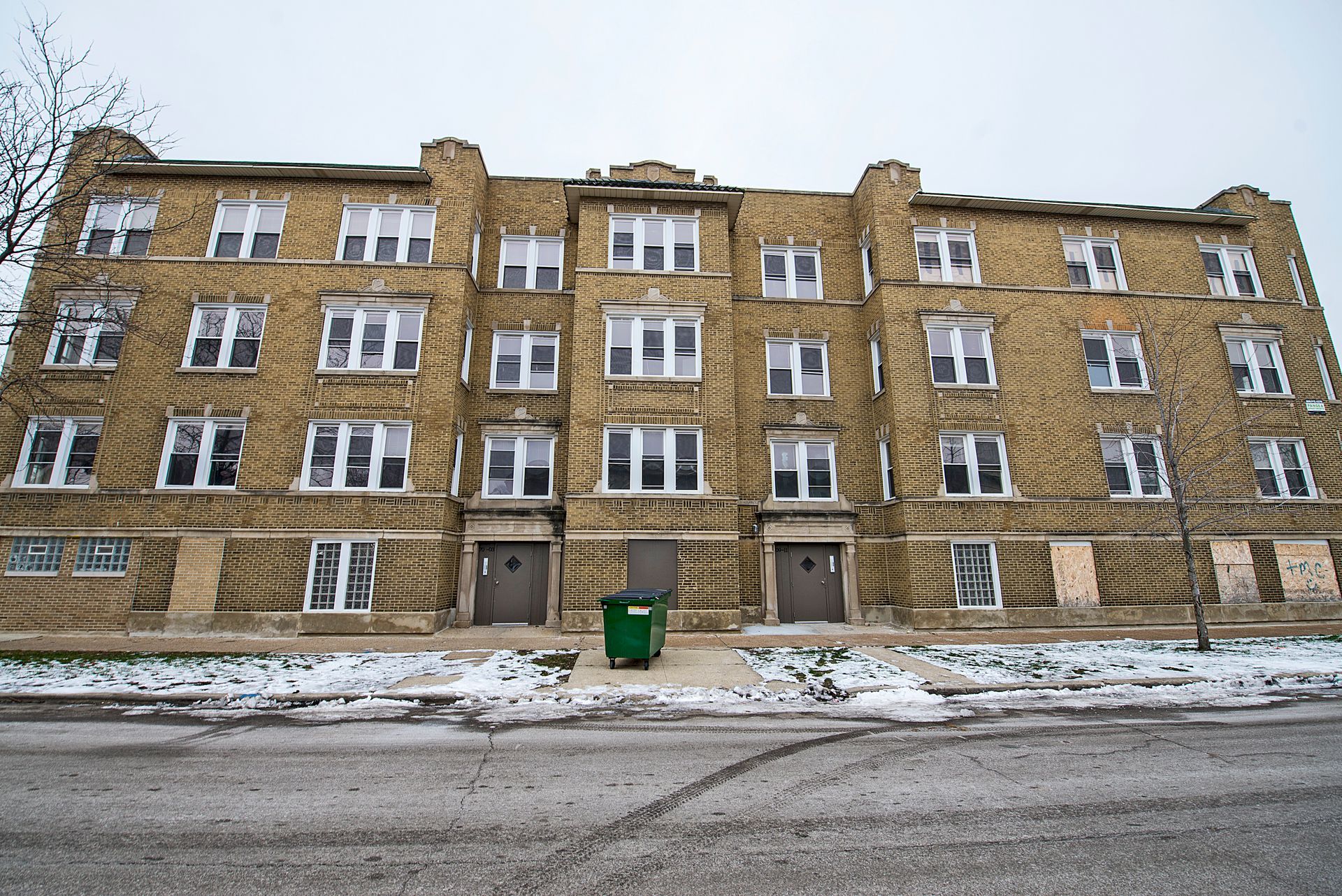 Three-story beige apartment building with multiple windows. Green trash bin sits between two doors. Snow on ground.