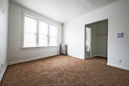 Empty room with brown carpet, window, white walls, and an open doorway to a bathroom.