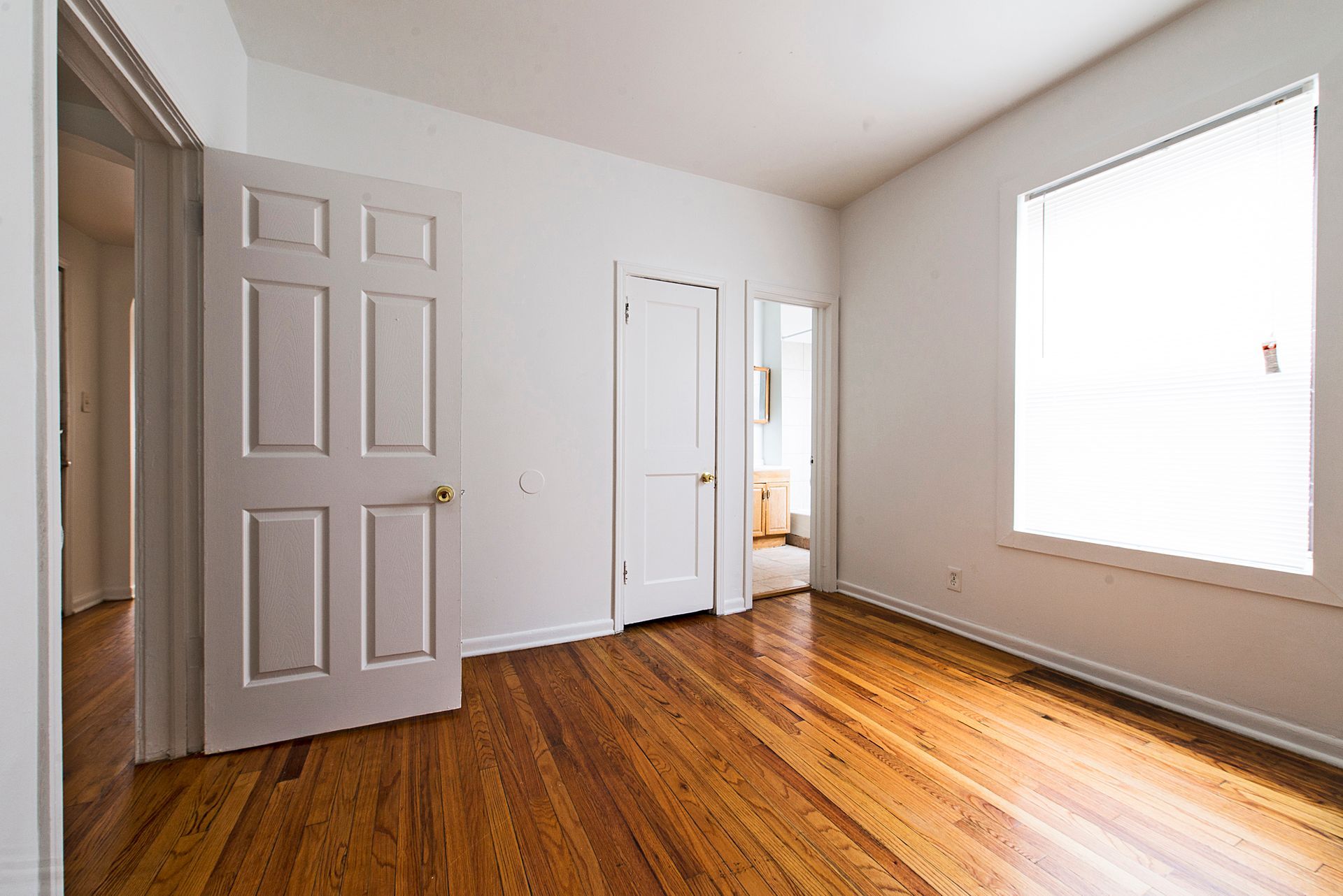Empty room with hardwood floors, white walls, doors, and a window with blinds.