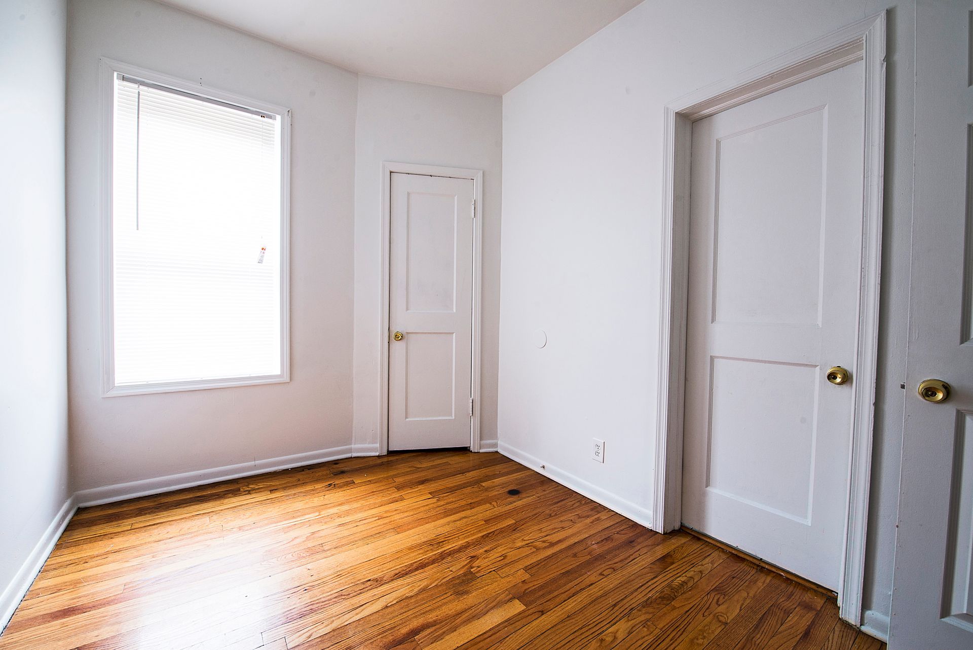 Empty room with hardwood floors, white walls, two doors, and a window with blinds.