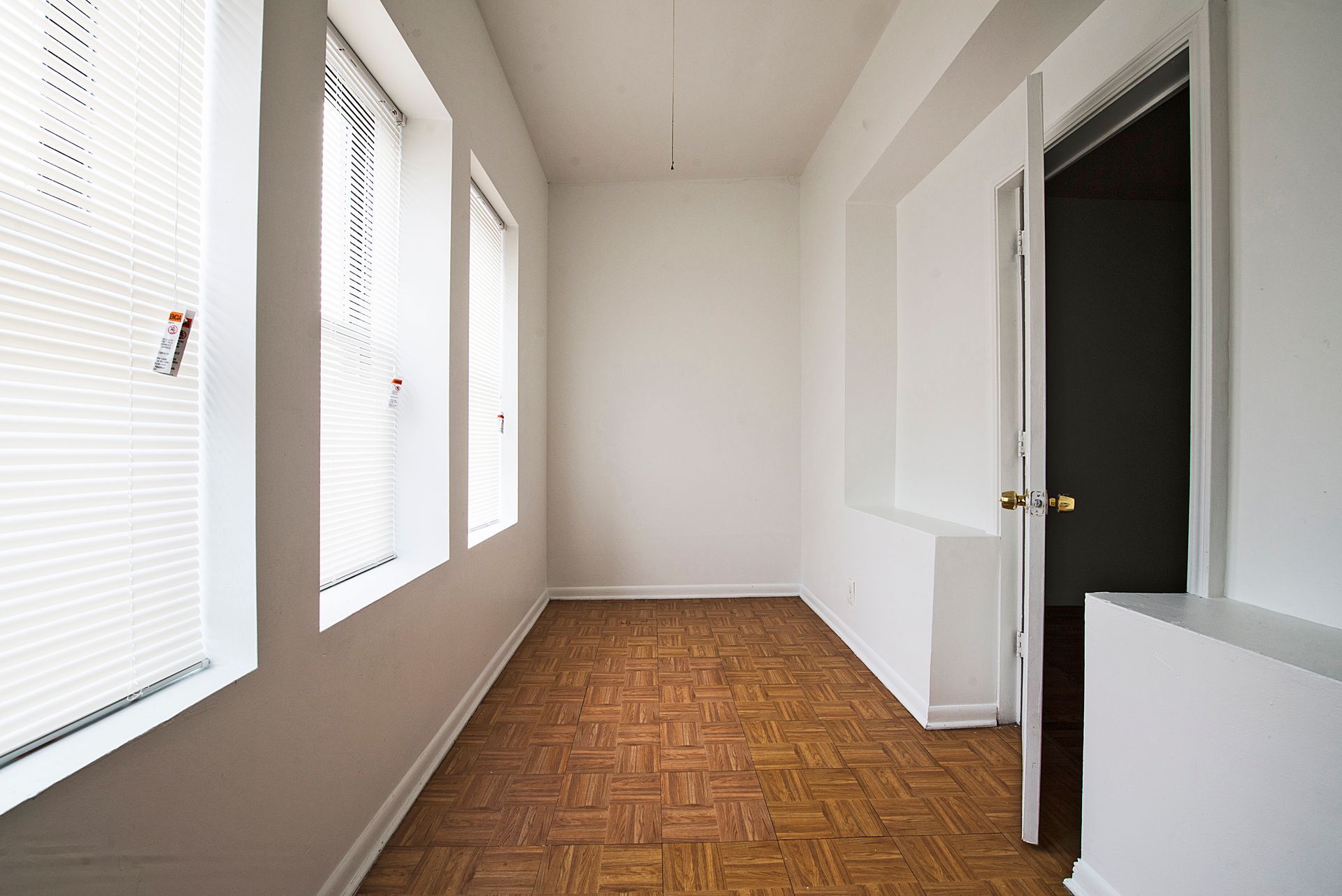 Narrow, empty room with windows and open doorway; wood-look flooring; white walls.