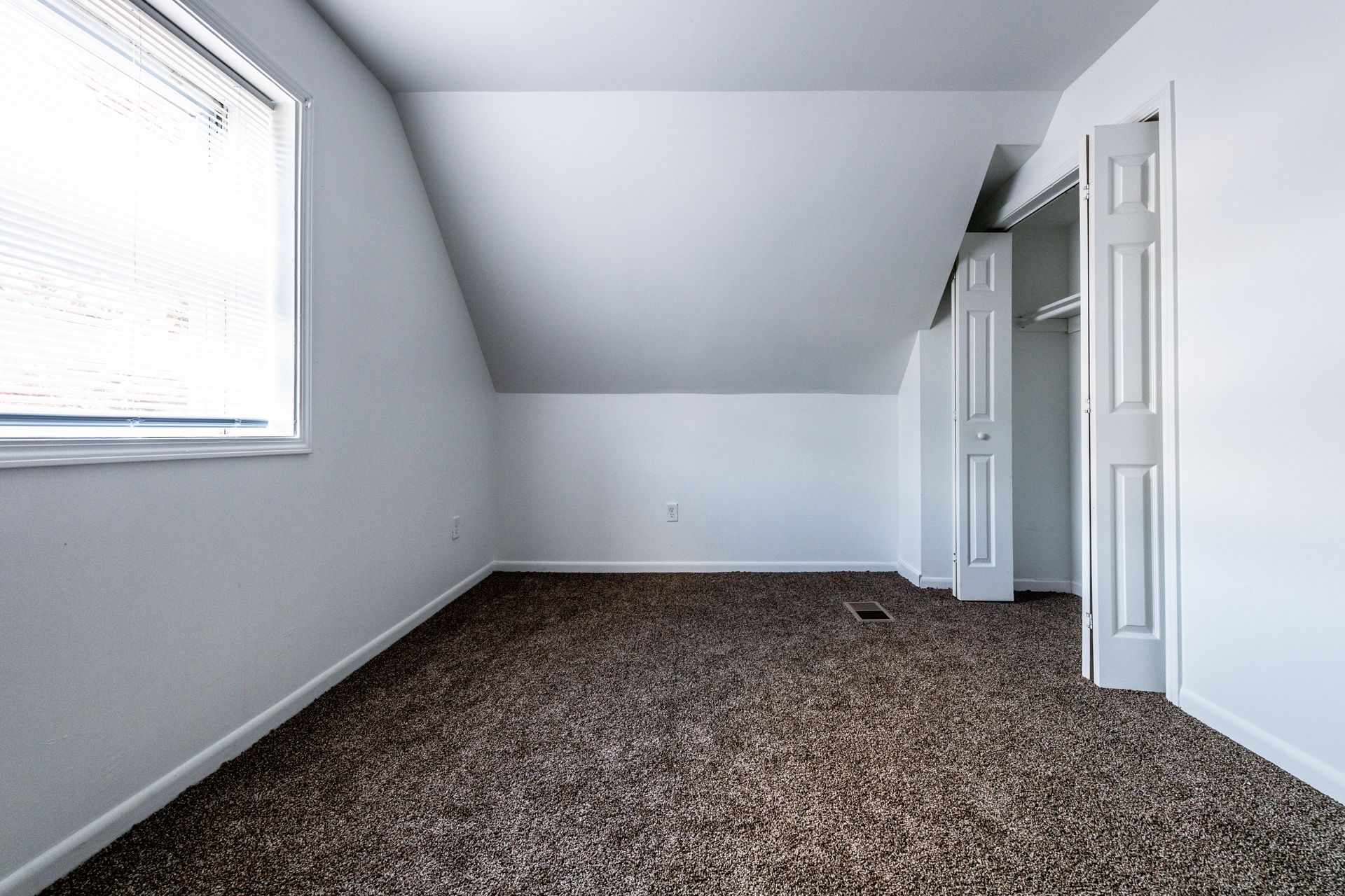 Empty, white-walled room with sloped ceiling, window, and closet doors. Brown carpet covers the floor.