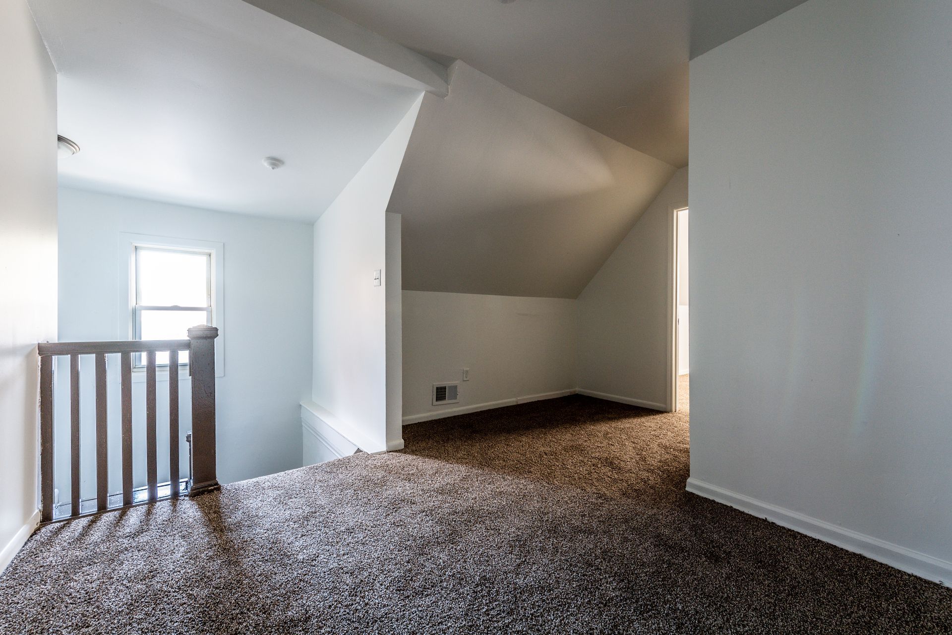 Interior view of a carpeted room with angled ceiling and window. A wooden railing is visible on the left.