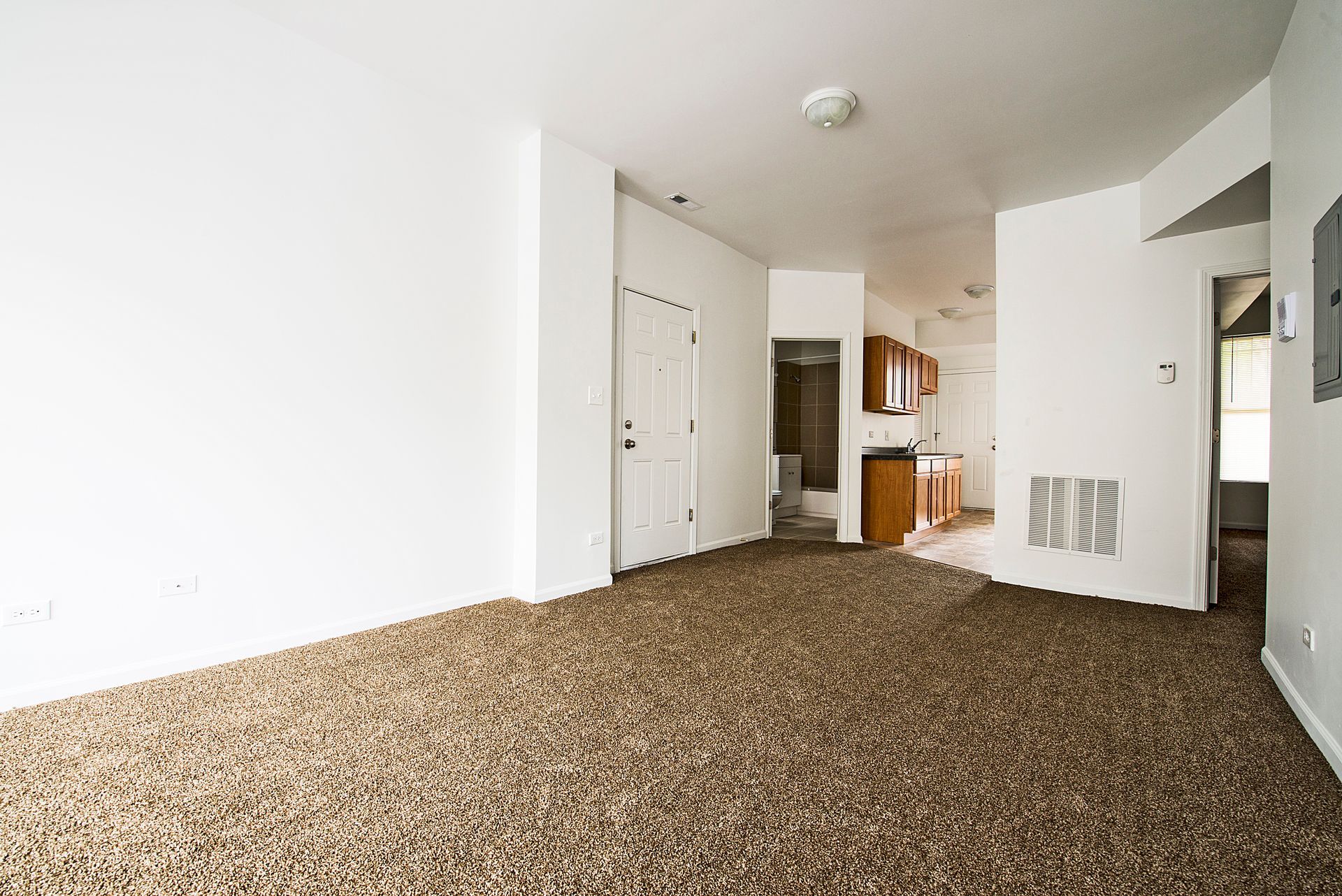 Empty room with brown speckled carpet, white walls, and doorway to kitchen.