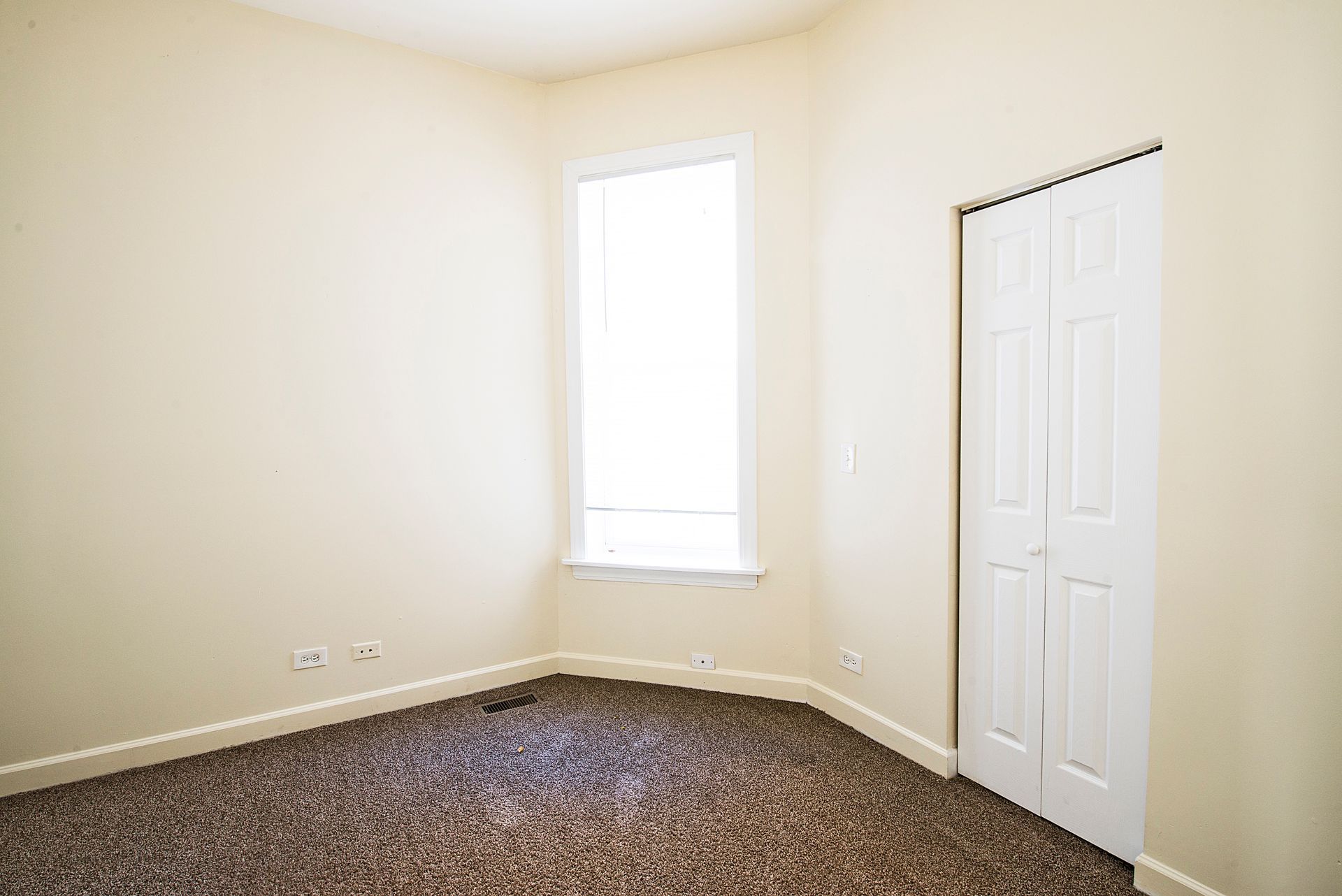 Empty room with beige walls, window, white door, and brown carpet.