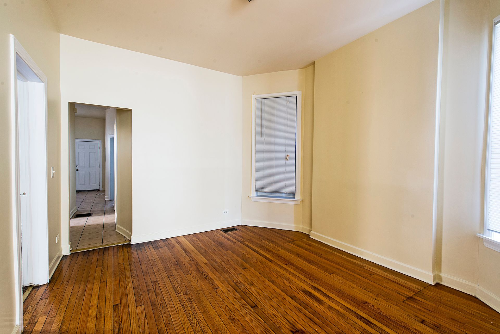 Empty room with hardwood floors, beige walls, and window. Doorway leads to another room.