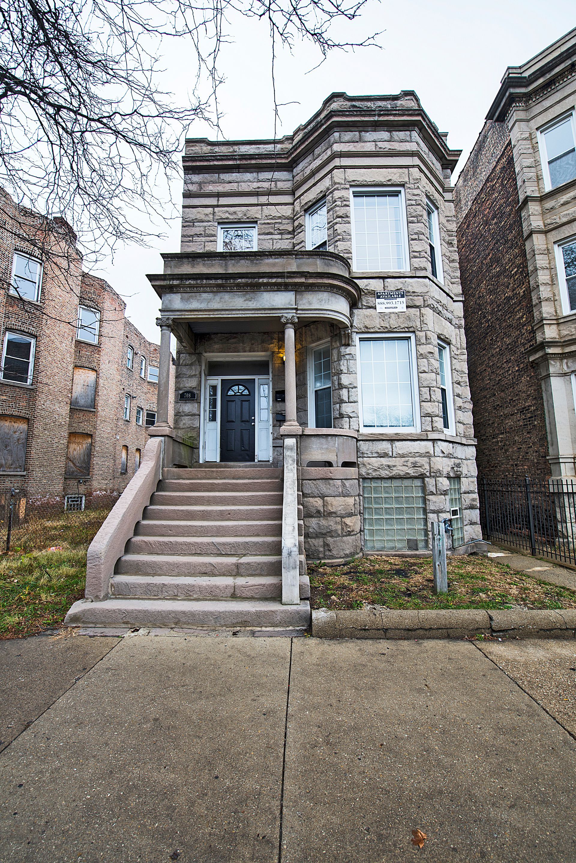 Three-story stone building with steps leading to the front door. Grey sky, sidewalk, and adjacent brick building.