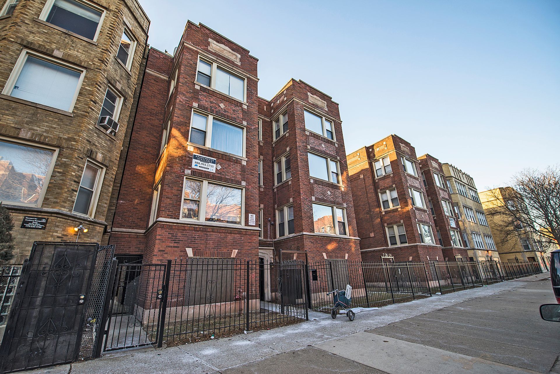 Brick apartment buildings on a city street, partially fenced off, with cloudy sky.