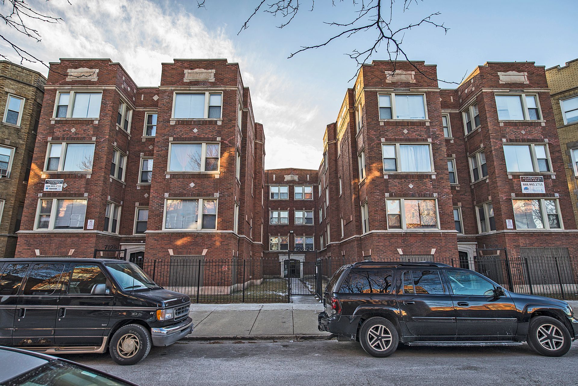 Red brick apartment buildings with parked black vehicles in front.