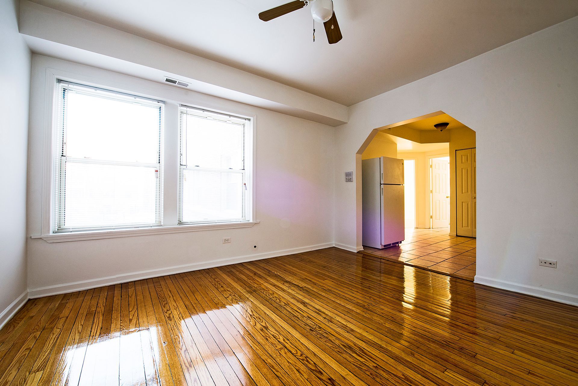 Empty room with hardwood floors, large windows, and an arched doorway leading to a kitchen.