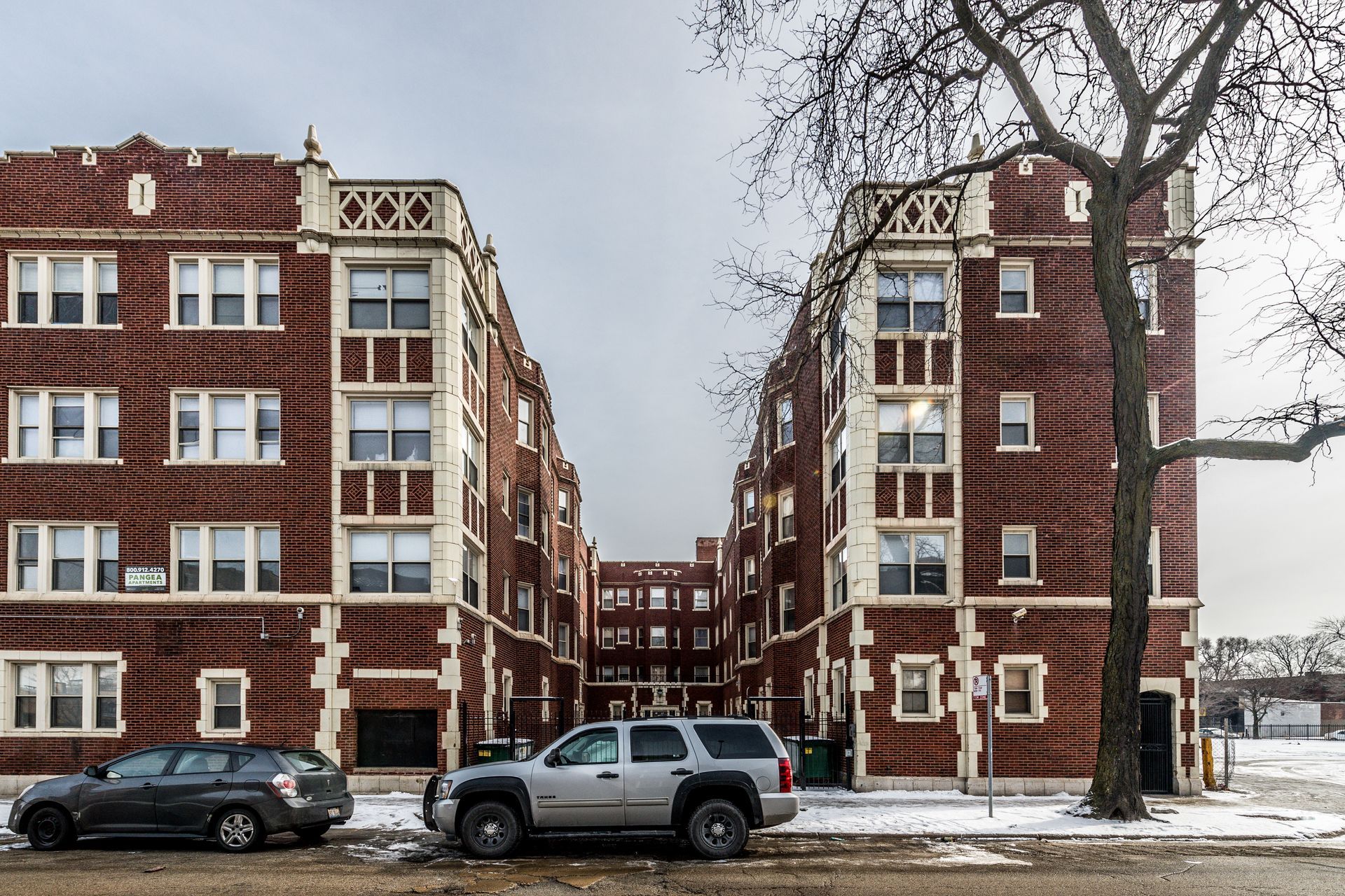 Two brown brick apartment buildings, a tree, and cars on a snowy street.