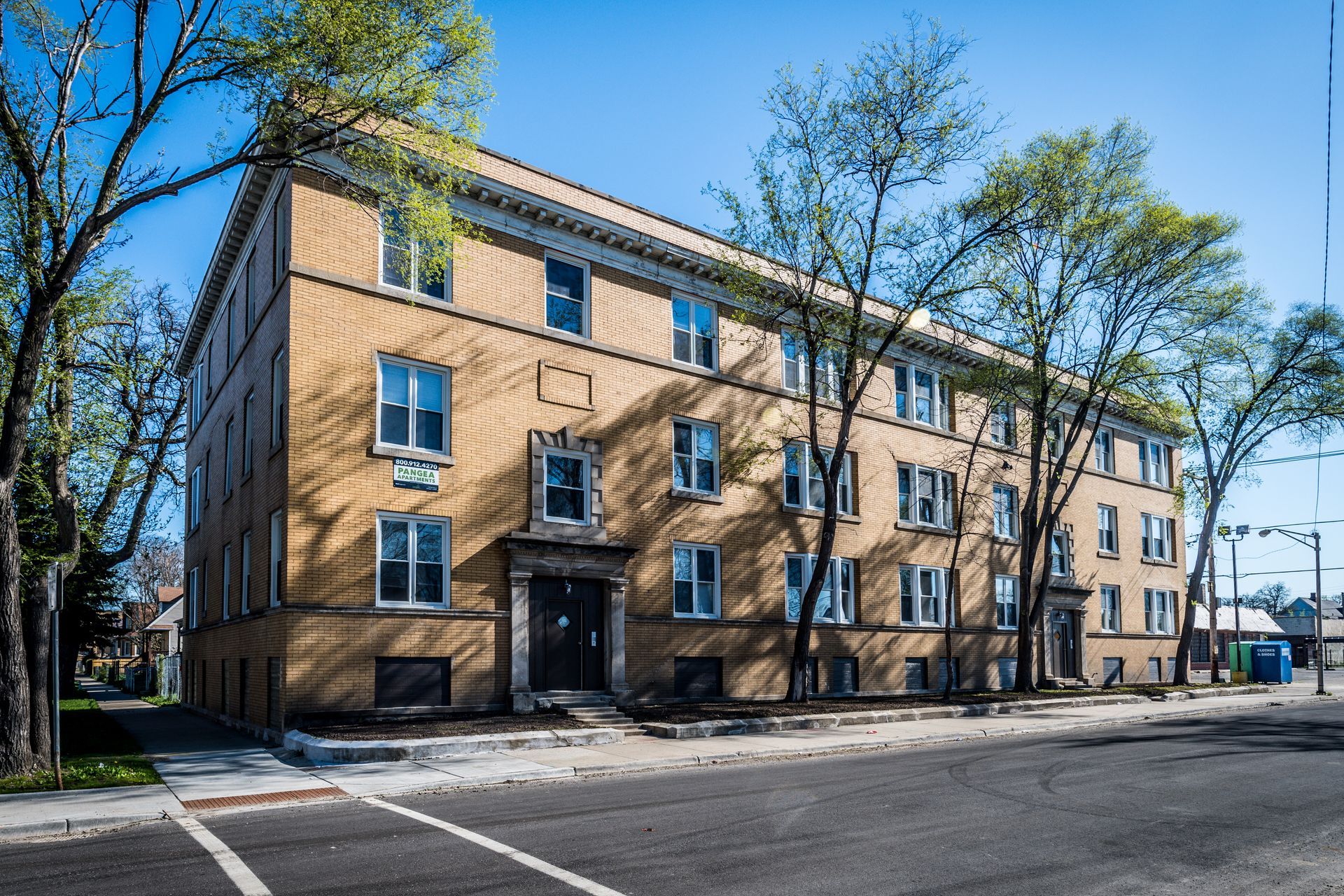 Three-story brick apartment building on a city street, trees in the foreground, blue sky.