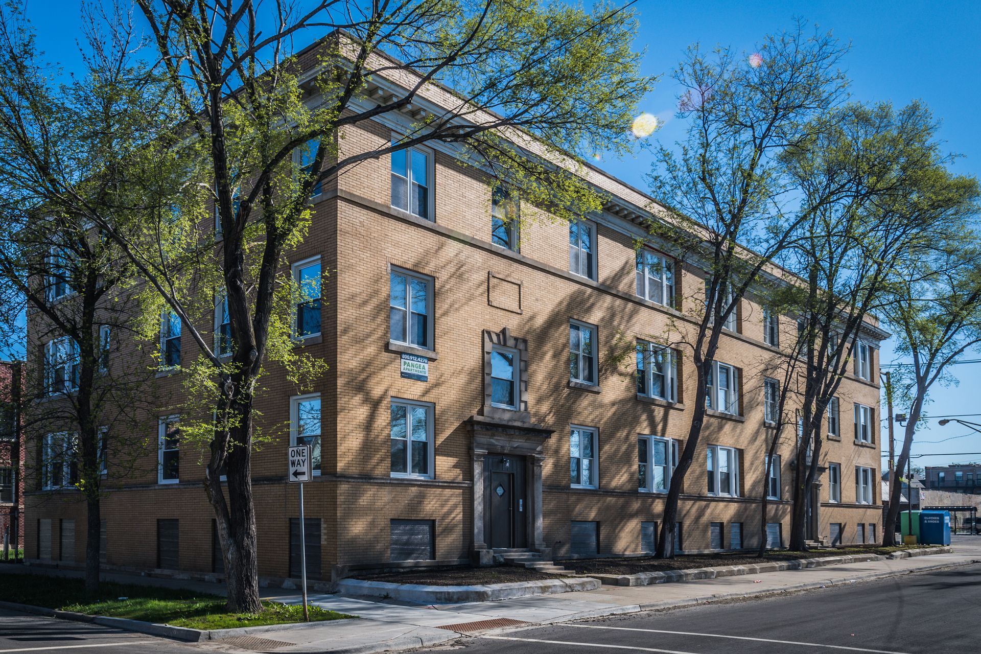 Three-story brick apartment building with multiple windows and trees in front on a sunny day.