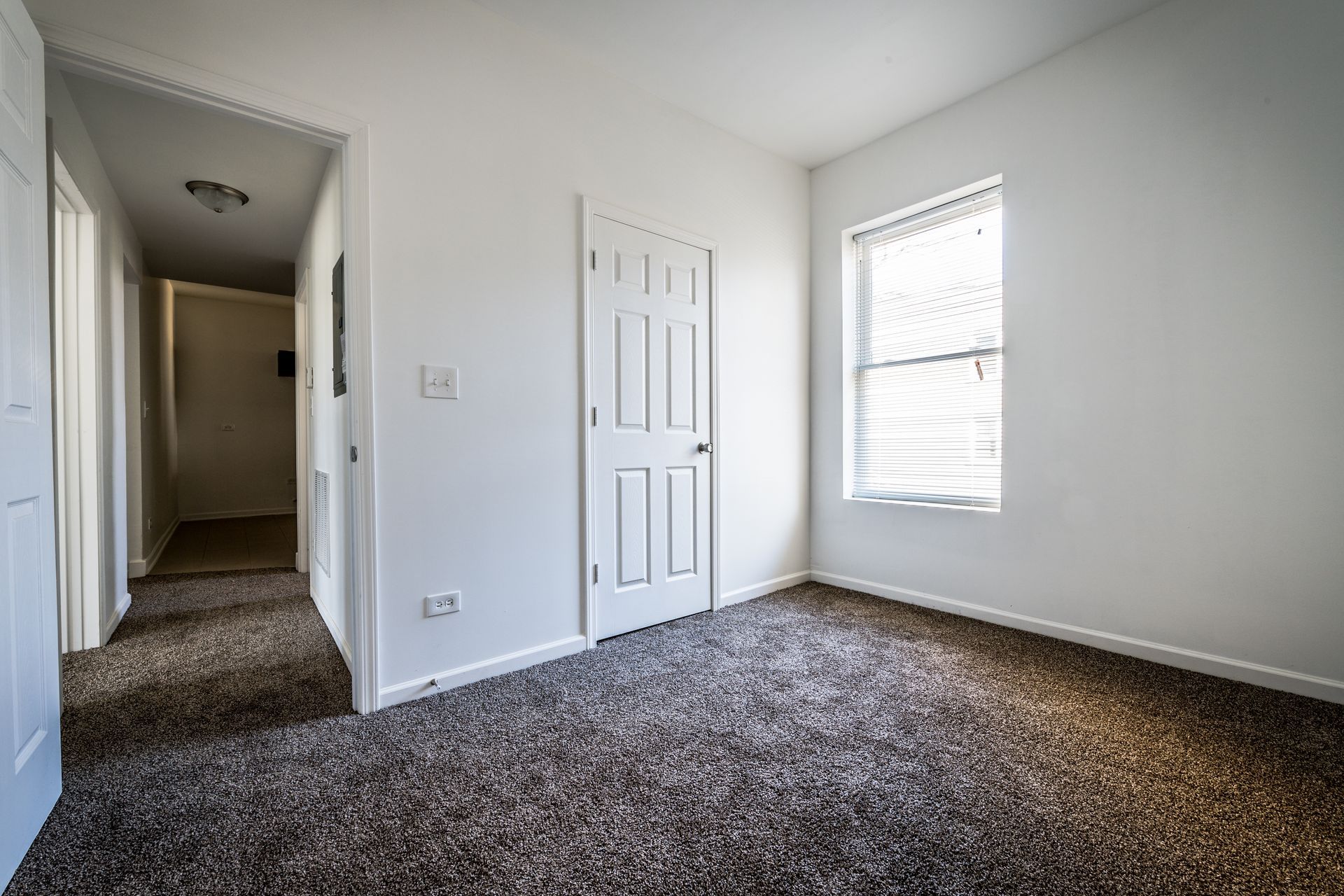 Empty bedroom with white walls, door, window, and brown carpet.