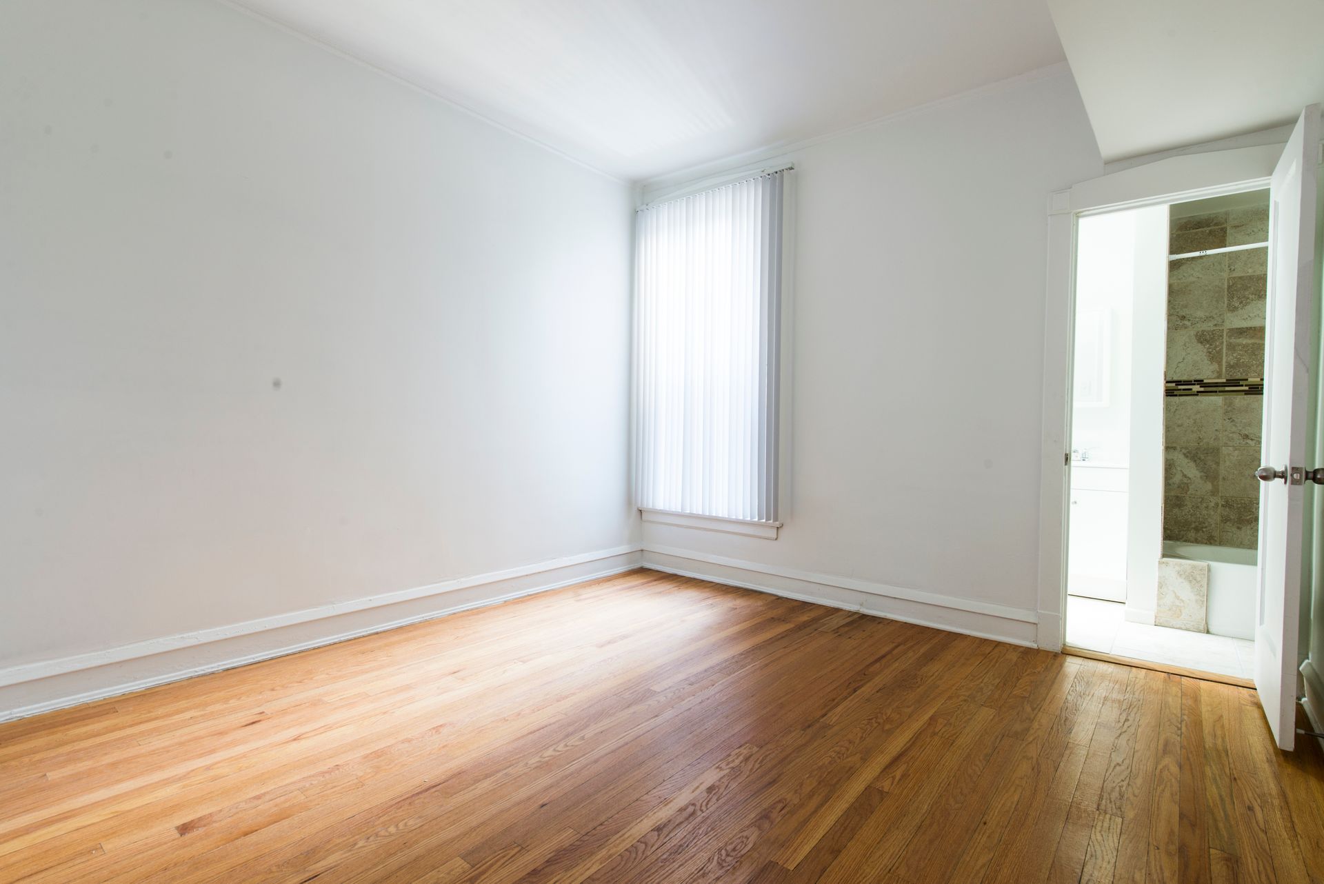 Empty room with hardwood floors, white walls, and a window with a sheer curtain; a doorway leads to a bathroom.