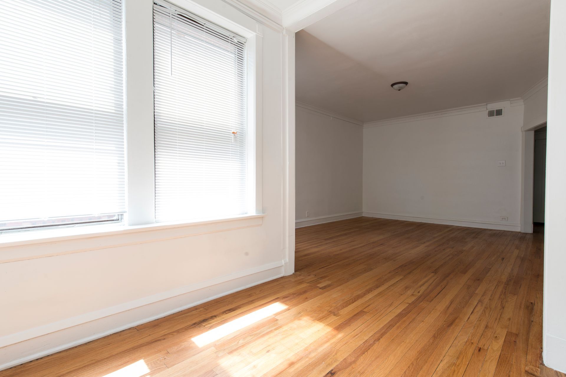 Empty room with hardwood floors, large window with blinds, and white walls.