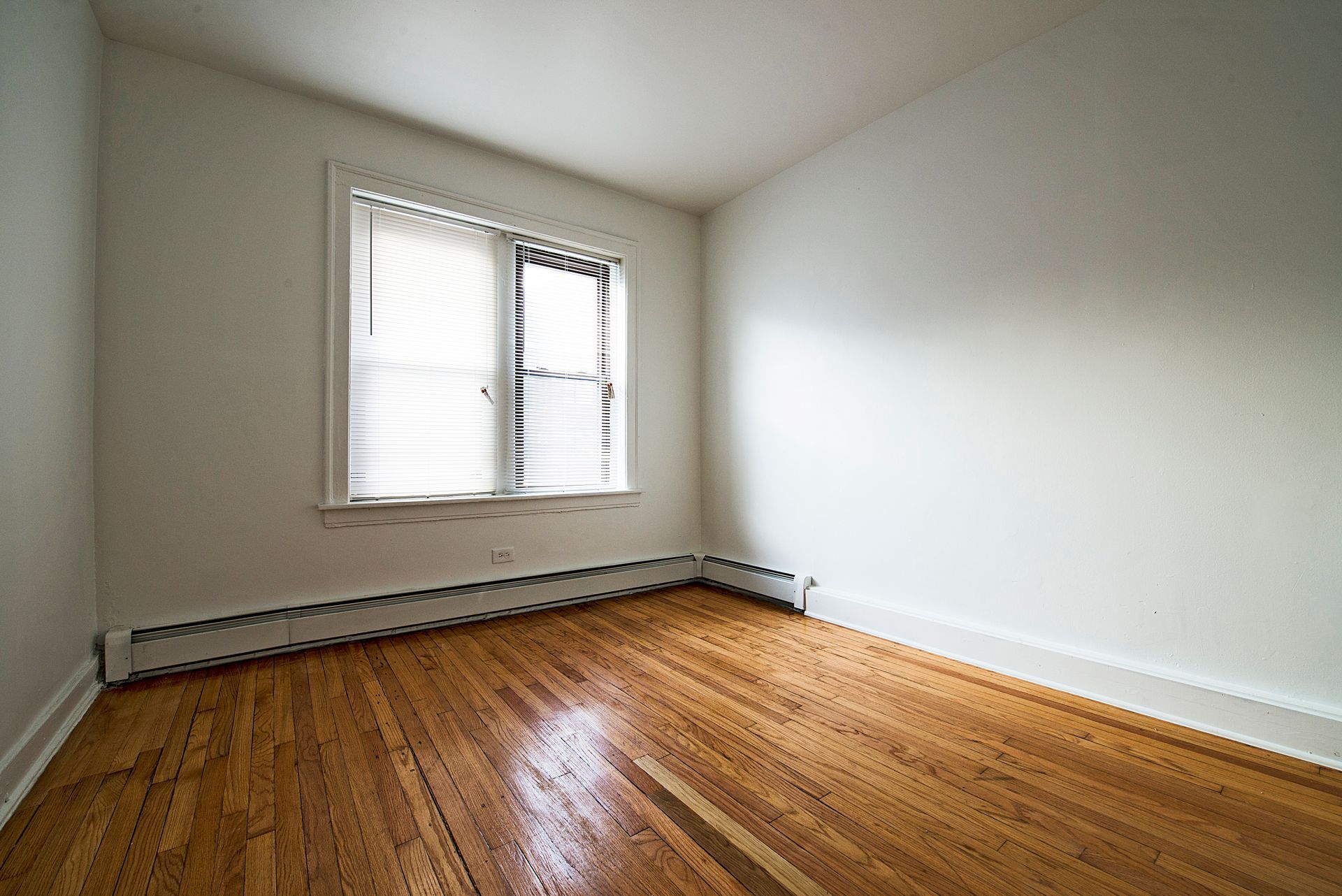 Empty room with hardwood floors, a window with blinds, and white walls.