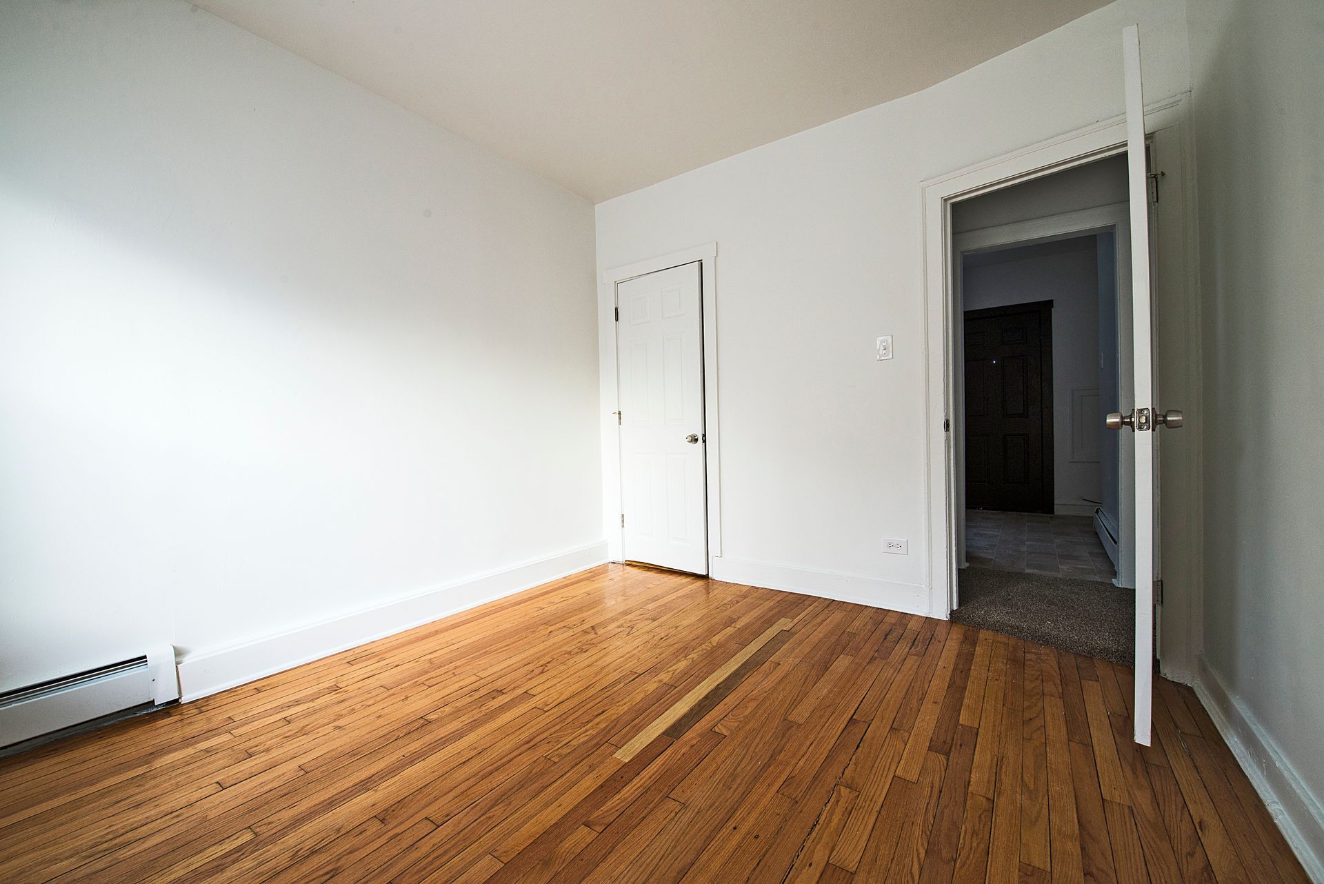 Empty room with hardwood floors, white walls, and a closed closet door. An open doorway leads to another room.