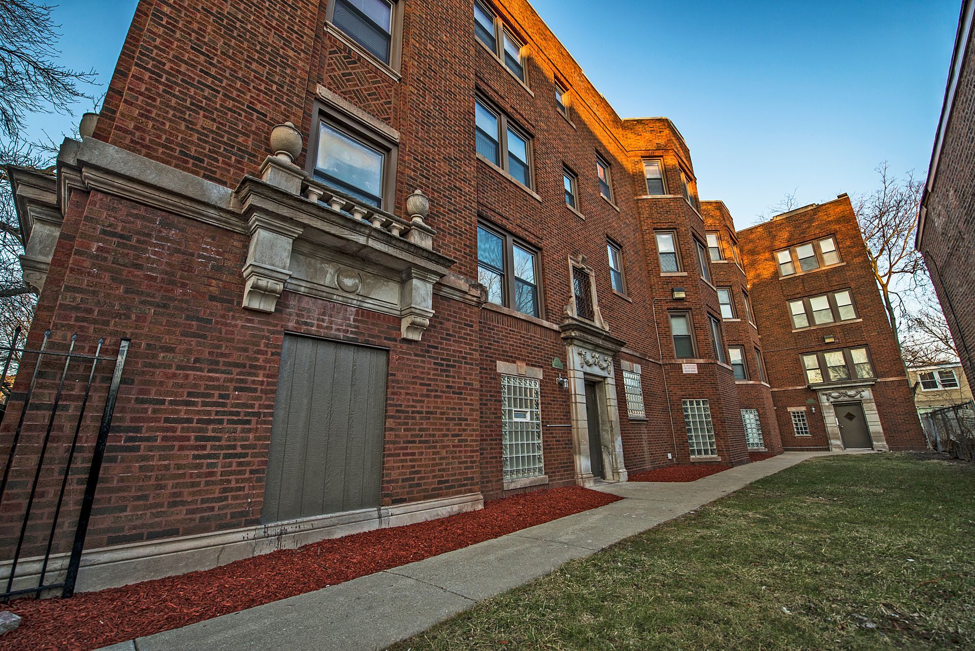 Red brick apartment building with architectural details; sidewalk and small lawn in front.