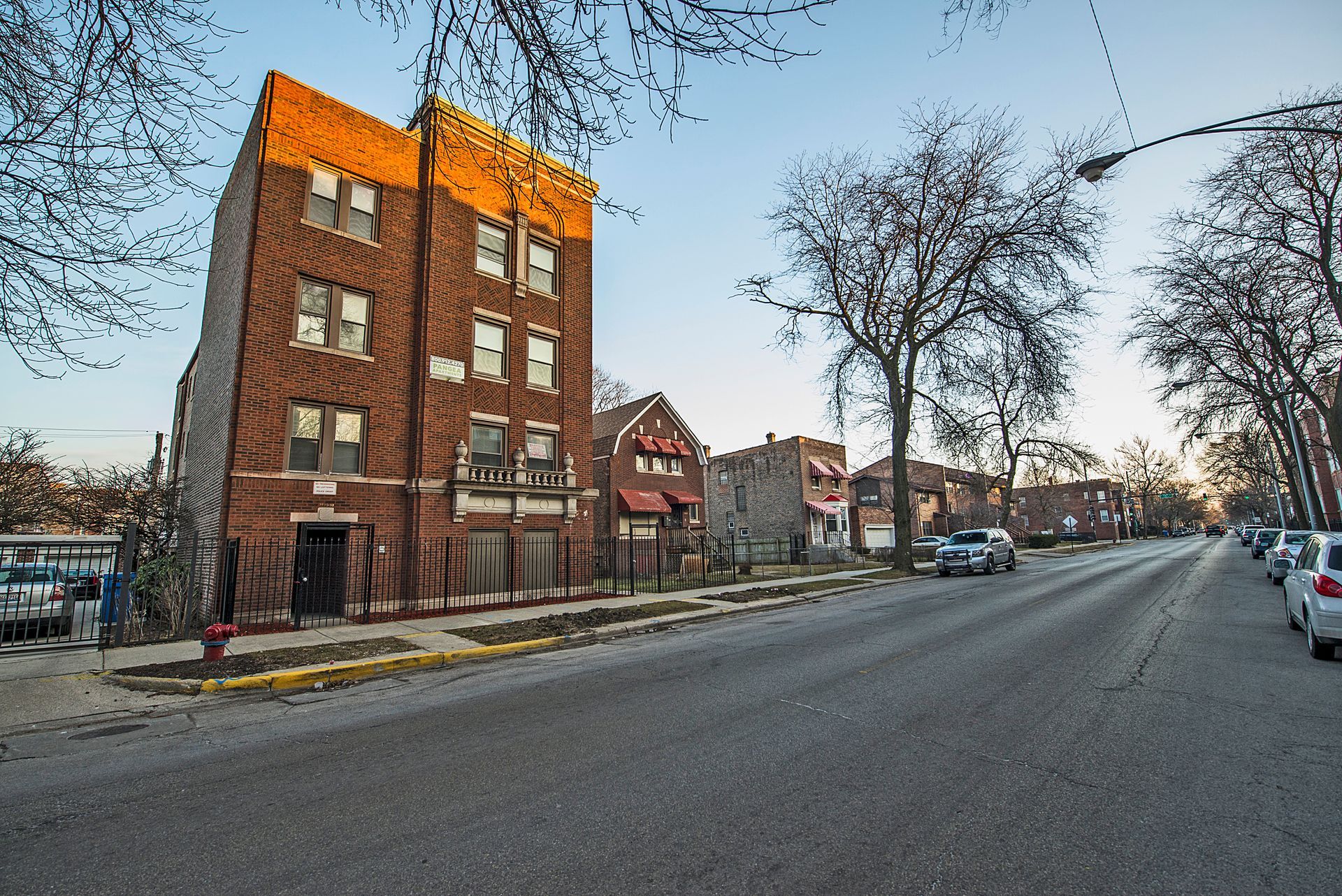 Brick apartment building with sunlit top, along a street with bare trees and parked cars.